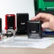 Hand greift nach einem Trodat-Stempel auf einem Schreibtisch mit Dokument und Brille