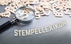 Nahaufnahme einer Lupe, die über verstreute hölzerne Buchstaben 'LEXIKON' fokussiert, mit 'STEMPELLEXIKON' Text auf grauem Hintergrund.